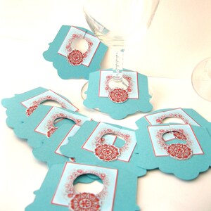 May include: Set of teal blue and red wine glass charms with a decorative design. The charms have a cut-out circle in the center for the stem of the glass.