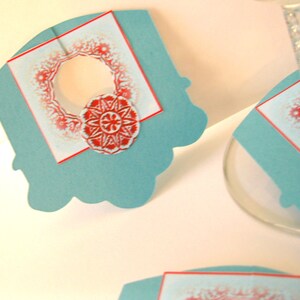 May include: Teal blue paper place card with a red and white snowflake design. The place card has a cut-out circle in the center.