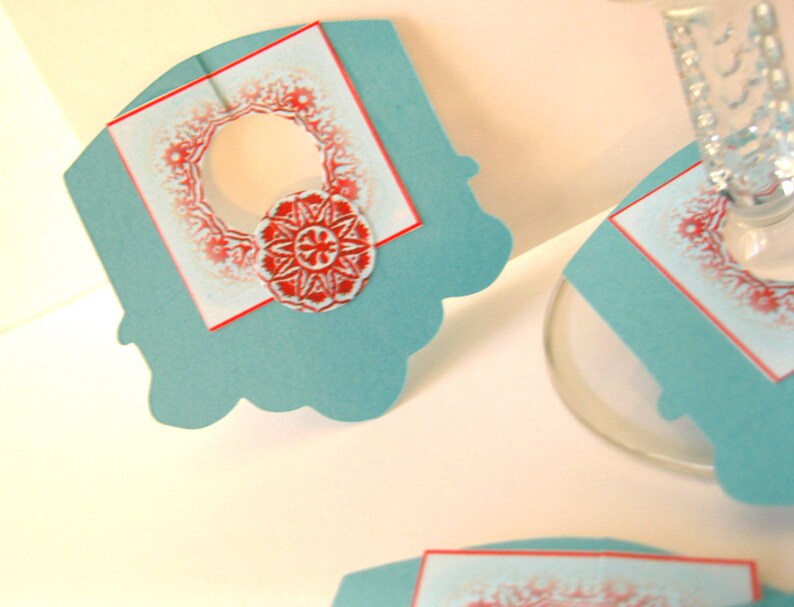 May include: Teal blue paper place card with a red and white snowflake design. The place card has a cut-out circle in the center.
