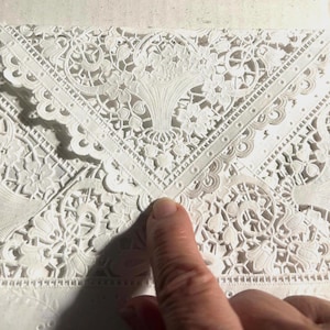 May include: Close-up of a white, intricately designed lace envelope. The lace features floral and geometric patterns, with a central floral motif. The word "ROYAL" is visible on the right side. A finger points to the envelope.
