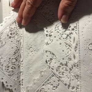 May include: Close-up of white, intricately designed paper lace. The lace features floral and geometric patterns, with delicate cut-out details. The paper is folded, revealing the detailed design. The image is well-lit, highlighting the texture and craftsmanship of the lace.