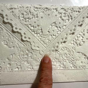 May include: A white, intricately designed envelope with a delicate lace-like pattern. The envelope features floral and geometric motifs, with scalloped edges and a rectangular area for text. The design is embossed, creating a textured surface.