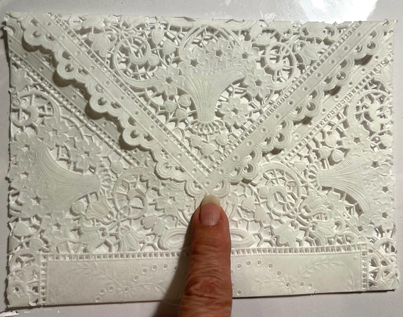 May include: A white, intricately designed envelope with a delicate lace-like pattern. The envelope features floral and geometric motifs, with scalloped edges and a rectangular area for text. The design is embossed, creating a textured surface.