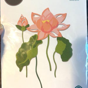 May include: A Sizzix Thinlits die set by Lisa Jones featuring 16 metal cutting dies to create a layered lotus flower, a bud, and lily pads. The packaging displays a pink lotus flower, a bud, and green leaves with the text 16 DIES.