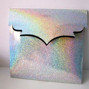 May include: A square, iridescent gift bag with a glittery finish. The bag has a decorative, black, curved cutout at the top. The bag's surface reflects a rainbow of colors, creating a shimmering effect.