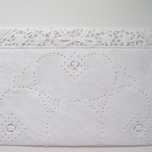 May include: A white, rectangular textile with intricate, raised floral and geometric patterns. The top edge features a delicate, openwork lace design with floral and bow motifs. The overall design is symmetrical and detailed, creating a vintage aesthetic.