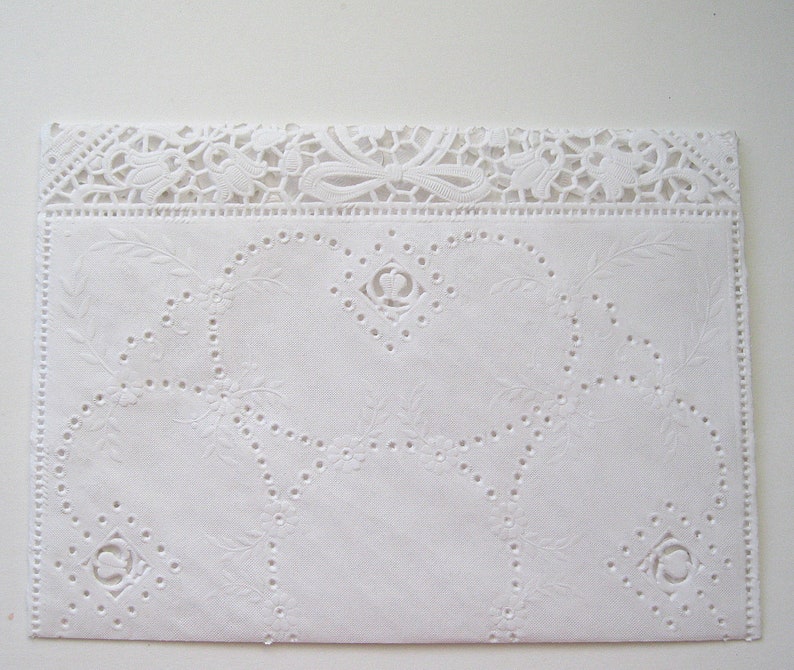 May include: A white, rectangular textile with intricate, raised floral and geometric patterns. The top edge features a delicate, openwork lace design with floral and bow motifs. The overall design is symmetrical and detailed, creating a vintage aesthetic.