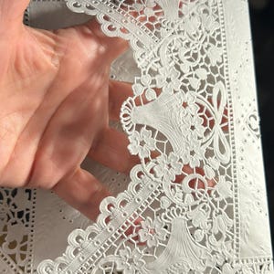 May include: White laser-cut paper with intricate floral and scrollwork designs. The paper features a delicate, openwork pattern with detailed edges and decorative elements, creating a lace-like appearance. The design includes stylized floral motifs and symmetrical arrangements.