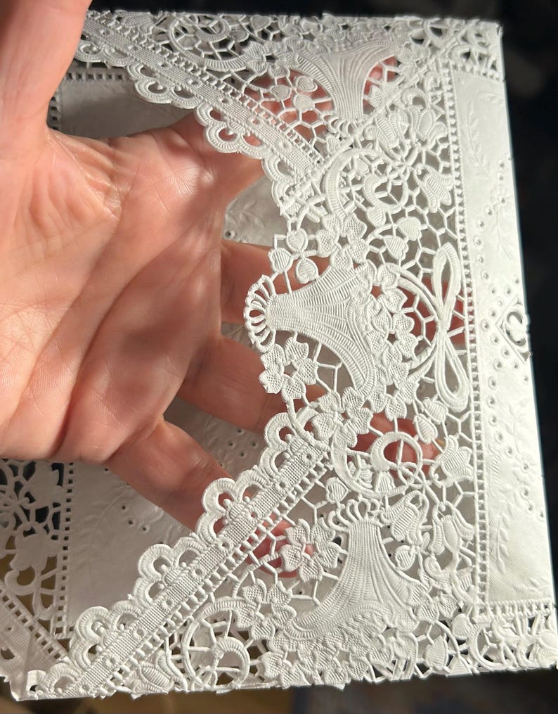 May include: White laser-cut paper with intricate floral and scrollwork designs. The paper features a delicate, openwork pattern with detailed edges and decorative elements, creating a lace-like appearance. The design includes stylized floral motifs and symmetrical arrangements.