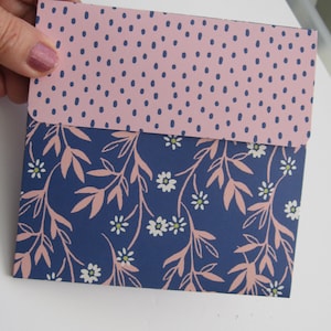 May include: A small, square envelope with a pink dotted flap and a navy blue floral design on the front. The floral pattern includes white flowers and peach-colored leaves. The envelope is held by a hand.