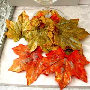 Puede incluir: Una colecci&oacute;n de hojas de arce decorativas de oto&ntilde;o en tonos naranja, rojo, dorado y verde. Las hojas est&aacute;n hechas de un material met&aacute;lico y tienen una superficie texturizada.