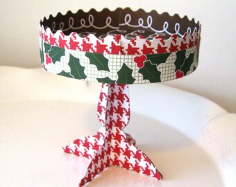Puede incluir: Un soporte para tartas de papel con un patrón rojo y blanco, un borde festoneado y un diseño de acebo y bayas. El soporte es perfecto para exhibir tartas o postres pequeños.