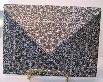 May include: A decorative envelope with a black and white floral pattern. The envelope is rectangular with a pointed flap. The design features swirling floral motifs. The envelope is displayed on a gold-colored wire stand.