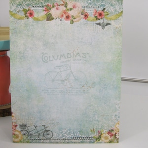 May include: A vintage-style paper with a faded blue background and a floral border. The paper features a black and white illustration of a bicycle with the text "COLUMBIAS HIGHEST GRADE ONLY" and "POPE MFG. CO., N. Y.  BOSTON, MASS.  CHICAGO, ILL.  SAN FRANCISCO, CAL.  TORONTO, CAN.  LONDON, ENG."