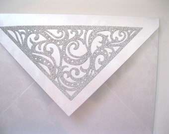 May include: A white envelope with a silver glitter-covered, triangular flap. The flap features a decorative, swirling, cut-out design. The envelope is a light, pearlescent color, suitable for invitations or special announcements.