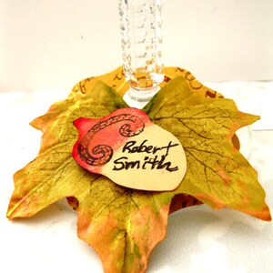 Puede incluir: Un charm para copa de vino con tem&aacute;tica oto&ntilde;al que presenta una hoja artificial con una bellota pintada y una etiqueta con el nombre "Robert Smith".