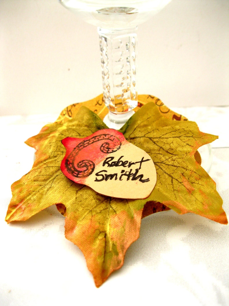 Puede incluir: Un charm para copa de vino con tem&aacute;tica oto&ntilde;al que presenta una hoja artificial con una bellota pintada y una etiqueta con el nombre "Robert Smith".