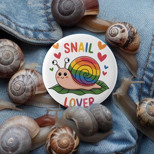 May include: A white button with a rainbow snail and the text "Snail Lover" surrounded by hearts. The button is on a blue denim background with several brown snail shells.