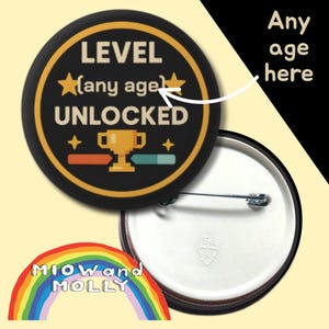 Op de afbeelding: Een zwarte button met de tekst "LEVEL (any age) UNLOCKED" in wit en goud. De button heeft een gouden trofee en een voortgangsbalk. De button is rond en heeft een speld op de achterkant.