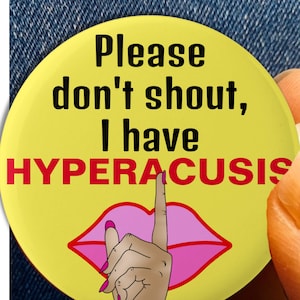 May include: Yellow button with the text "Please don't shout, I have HYPERACUSIS" in black and red. A pink finger with a pink fingernail is over the text, making a shushing gesture.