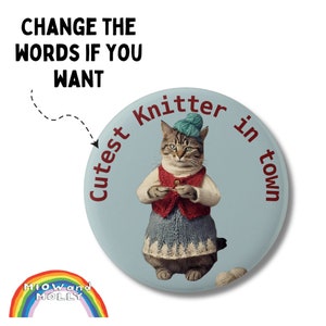 May include: A round, light blue button featuring a cat dressed as a knitter. The cat wears a red vest, blue skirt, and a green hat. Text around the cat reads "Cutest Knitter in town". A rainbow logo is in the bottom left.