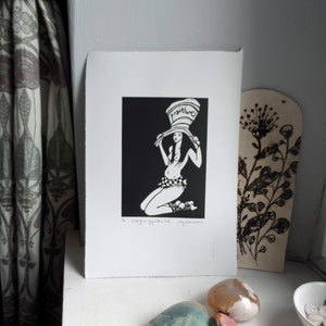 May include: A black and white print featuring a kneeling figure wearing a hat with the word "positive". The print is on white paper with a deckled edge. Decorative items are in the background.
