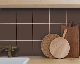 May include: A kitchen backsplash featuring square dark brown tiles with thin white grout lines. The wall is positioned behind a light wood countertop, a white sink, a brass faucet, and a wooden shelf holding white plates and glassware.