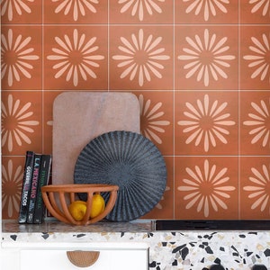 Puede incluir: Una encimera de cocina con un salpicadero de terracota con un patrón repetitivo de flores estilizadas blancas. La encimera está hecha de terrazo y tiene un armario blanco debajo. Un salvamanteles de pizarra negra y un cuenco de terracota con limones están sobre la encimera.