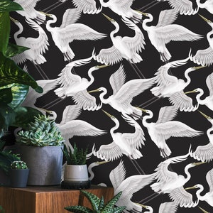 Op de afbeelding: Een zwart-wit behang met een herhalend patroon van witte reigers met uitgespreide vleugels. De reigers vliegen in een diagonaal patroon tegen een zwarte achtergrond.