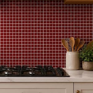May include: A kitchen scene featuring a black gas cooktop set into a white countertop against a deep red square-tiled backsplash. Above, a stainless steel range hood sits between two wooden shelves. A wooden cutting board and utensils are visible.