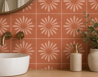May include: A bathroom wall featuring square terracotta tiles with a repeating light pink floral star pattern. A white ceramic vessel sink, a brass faucet, a beige soap dispenser, and a small potted plant are arranged on a wooden countertop.