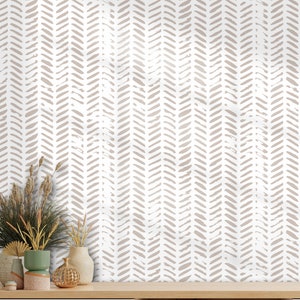 May include: A white shelf unit with a light wood finish. The back wall is covered in a beige and white chevron patterned wallpaper. The shelf unit has two drawers and an open shelf. There are decorative items on the top shelf, including a vase with dried flowers, a gold-colored decorative object, and a small potted plant on the bottom shelf.