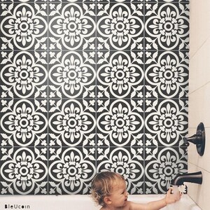 A baby in a bathtub playing with toy cars, with the wall behind the tub covered in black and white patterned tile stickers.
