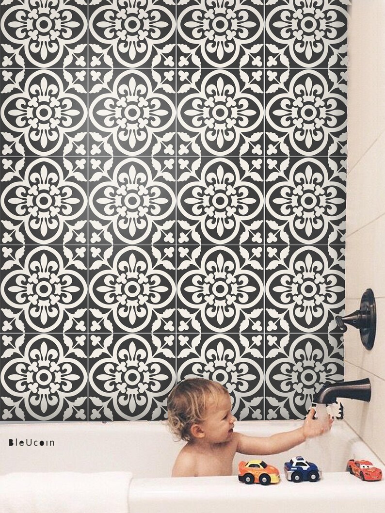 A baby in a bathtub playing with toy cars, with the wall behind the tub covered in black and white patterned tile stickers.