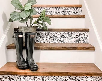 Könnte beinhalten: Eine Treppe mit dunklen Holzstufen und weißen Setzstufen, die mit einem komplexen schwarz-weißen Kreismuster verziert sind. Ein Paar schwarze Hunter-Gummistiefel und eine grüne Pflanze in einem Eimer aus verzinktem Metall stehen auf einer Stufe.
