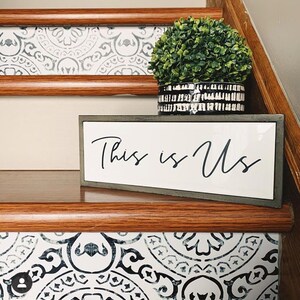 A close-up of a staircase riser with a black and white patterned decal, with a small potted plant and a "This is Us" sign on the wooden tread.