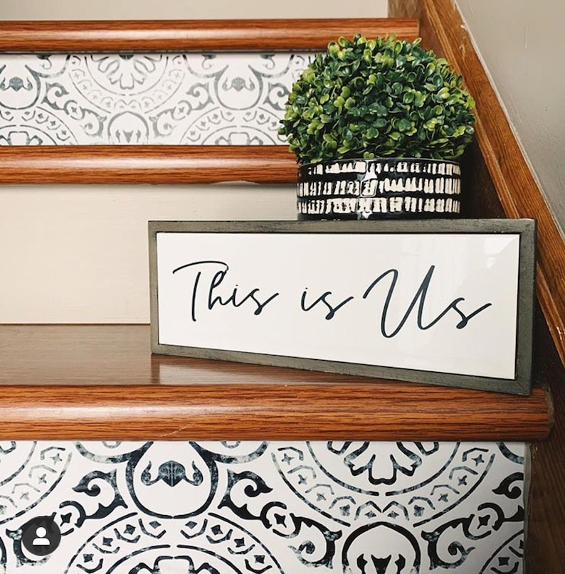 A close-up of a staircase riser with a black and white patterned decal, with a small potted plant and a "This is Us" sign on the wooden tread.