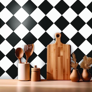 Op de afbeelding: Een houten snijplank, lepel, vork en klein potje staan op een houten oppervlak. Twee kleine vazen met gedroogde planten staan rechts. De achtergrond is een zwart-wit dambordpatroon.