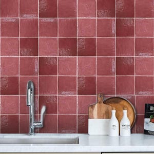 Könnte beinhalten: Ein Küchenrückwandspiegel mit quadratischen, glänzenden, dunkelroten Fliesen mit dezenter Textur. Ein silberner Wasserhahn steht links, während ein Holzschneidebrett, zwei weiße Flaschen mit der Aufschrift Öl und Essig sowie ein Buch auf der Arbeitsplatte stehen.