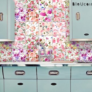 Op de afbeelding: Een keukenachterwand met een bloemmotief tegelontwerp. De tegels zijn wit met roze, rode en paarse bloemen. De achterwand bevindt zich achter een lichtblauw keukenblad met drie lades.
