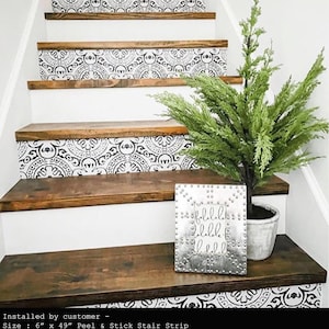 A staircase with dark wood treads and white risers covered in black and white patterned decals, with a small faux Christmas tree and a silver studded sign on the first step.