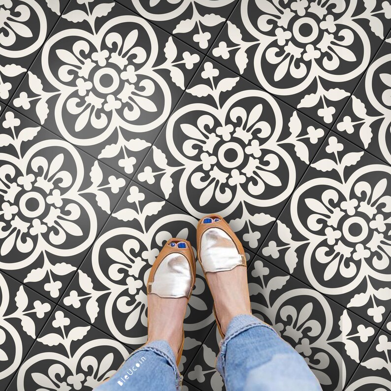 A person's feet in gold shoes and blue jeans standing on a floor covered in black and white patterned tile stickers.