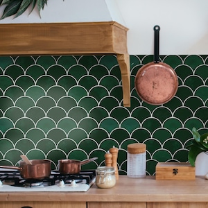 May include: A kitchen with a dark green, fish-scale tile backsplash. Copper pans are on the hob and hanging on the wall. Wooden cabinets and a wooden range hood complete the kitchen design, with a rustic feel.