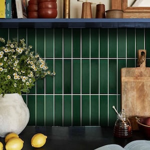 Può includere: Un paraschizzi da cucina con piastrelle rettangolari verde scuro disposte verticalmente con fughe bianche. La scena include un vaso bianco con margherite, due limoni gialli, un tagliere in legno e un vasetto di miele in vetro con mestolo.