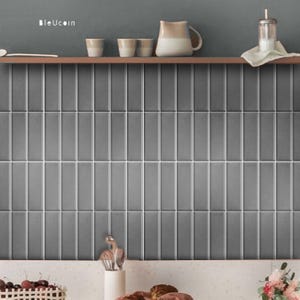 Può includere: Un rivestimento per cucina con piastrelle grigie rettangolari disposte verticalmente e fughe bianche. Sopra le piastrelle c'è una mensola in legno con tre tazzine, una brocca in ceramica e un barattolo di vetro su un panno bianco. Il testo BleUcoin è visibile.