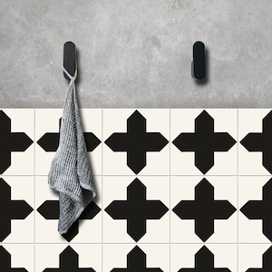 Puede incluir: Dos ganchos negros montados en una pared gris sobre un suelo de baldosas. El suelo está enlosado con cruces geométricas negras y blancas. Una toalla gris cuelga de uno de los ganchos.