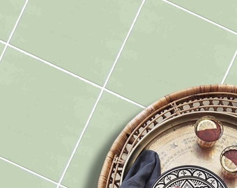May include: A round woven rattan tray holding a black and white patterned ceramic plate with fresh figs, two glasses with lemon slices, and a dark gray cloth. The tray is placed against a background of light sage green square tiles with white grout.