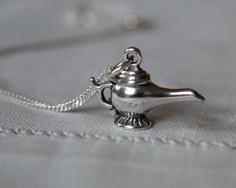 May include: A silver-colored pendant necklace featuring a detailed genie lamp charm. The charm has a classic design with a spout, handle, and ornate base. The necklace chain is also silver and visible against a soft, neutral background. The pendant is approximately 1 inch in height.