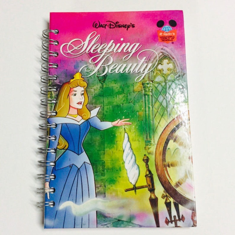 May include: A pink spiral bound book with a colourful illustration of Princess Aurora from Disney's Sleeping Beauty. The book cover features the title "Walt Disney's Sleeping Beauty" in a white cursive font.