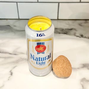 May include: A 16 fl oz can candle shaped like a beer can with the words "Natural Light" printed on the side. The candle is yellow and has a cork lid.